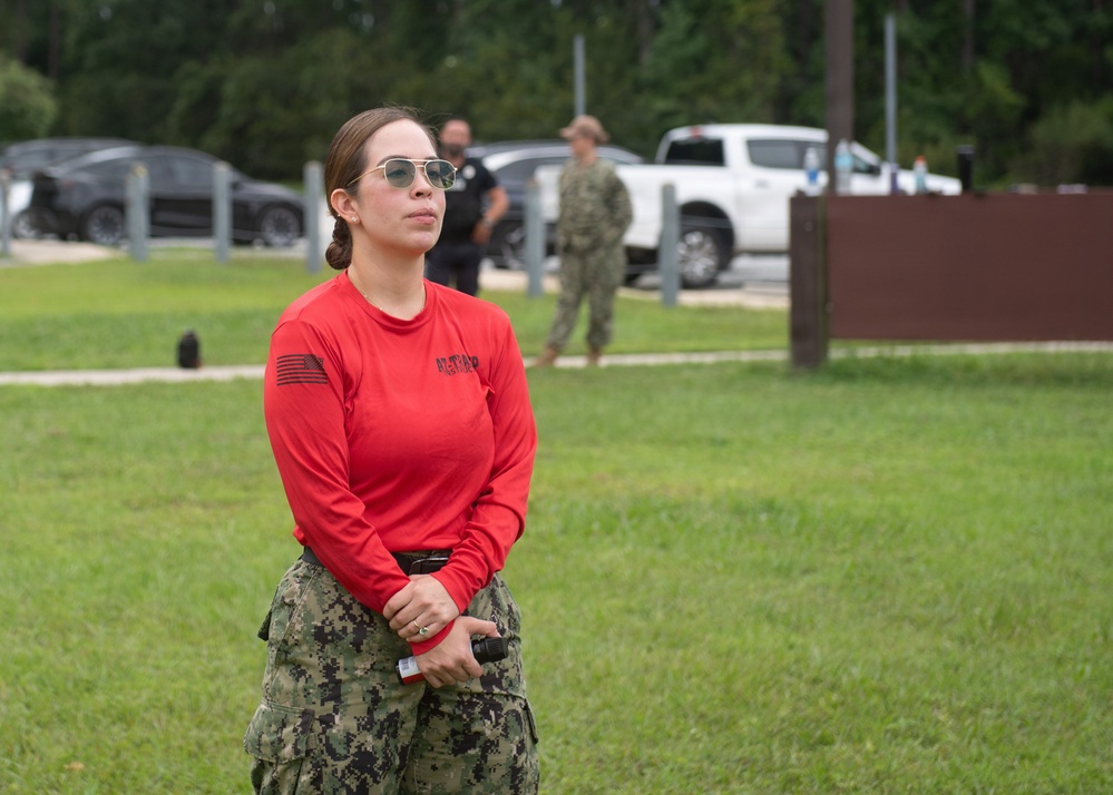 A female service member wearing a red long-sleeve shirt with a U.S. flag patch on the sleeve and digital camouflage (NWU Type I/Navy pattern) trousers stands on a grass field, holding what appears to be a small device or radio. She is wearing aviator-style sunglasses and has her hair pulled back. In the background, additional personnel in military and civilian attire are visible near parked vehicles.