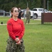 A female service member wearing a red long-sleeve shirt with a U.S. flag patch on the sleeve and digital camouflage (NWU Type I/Navy pattern) trousers stands on a grass field, holding what appears to be a small device or radio. She is wearing aviator-style sunglasses and has her hair pulled back. In the background, additional personnel in military and civilian attire are visible near parked vehicles.