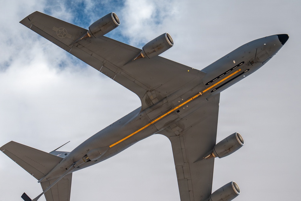 A bottom-up view of a large military jet aircraft, identifiable as a US Air Force tanker (likely a KC-135 Stratotanker), photographed from directly below while in flight. The aircraft displays a distinctive yellow/orange stripe along its fuselage underside and a US military star-and-bar insignia on the wing. Four pod-mounted jet engines are visible, consistent with the Boeing 707-derived airframe.