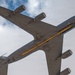 A bottom-up view of a large military jet aircraft, identifiable as a US Air Force tanker (likely a KC-135 Stratotanker), photographed from directly below while in flight. The aircraft displays a distinctive yellow/orange stripe along its fuselage underside and a US military star-and-bar insignia on the wing. Four pod-mounted jet engines are visible, consistent with the Boeing 707-derived airframe.
