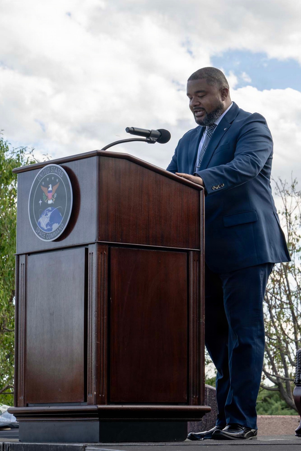 A man in a navy blue suit is speaking at an outdoor podium bearing the official seal of United States Space Command. The event appears to be a formal ceremony or official address, taking place under a partly cloudy sky with trees visible in the background. The speaker is looking downward, likely reading from prepared remarks.