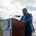 A man in a navy blue suit is speaking at an outdoor podium bearing the official seal of United States Space Command. The event appears to be a formal ceremony or official address, taking place under a partly cloudy sky with trees visible in the background. The speaker is looking downward, likely reading from prepared remarks.