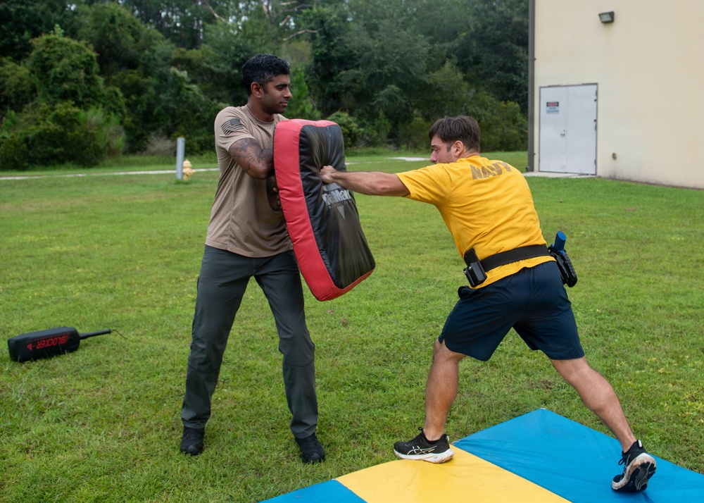 Two individuals are engaged in a combat training drill outdoors on a grass field near a building. One person in a tan shirt holds a large red and black striking pad while the other, wearing a yellow 'NAVY' shirt and a duty belt, delivers a strike to the pad. They are standing on or near blue and yellow training mats.