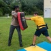 Two individuals are engaged in a combat training drill outdoors on a grass field near a building. One person in a tan shirt holds a large red and black striking pad while the other, wearing a yellow 'NAVY' shirt and a duty belt, delivers a strike to the pad. They are standing on or near blue and yellow training mats.