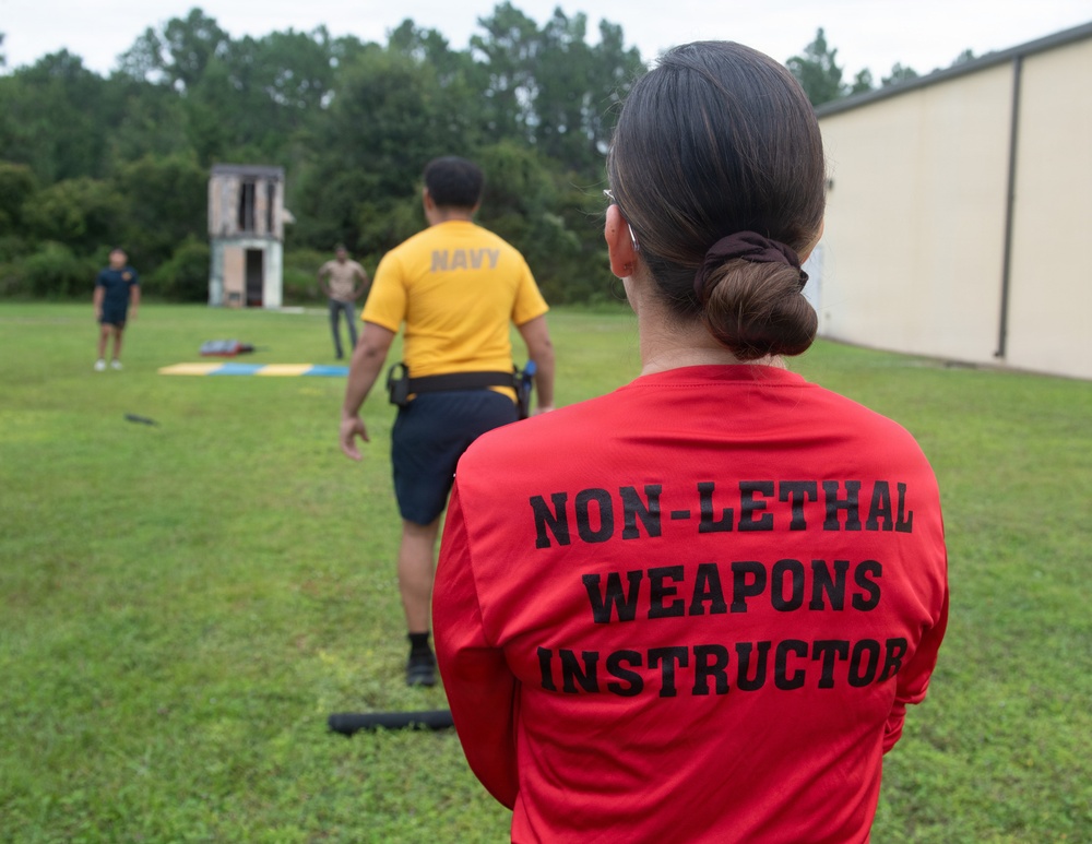 An outdoor military training exercise is taking place on a grassy field adjacent to a building. A person wearing a red shirt labeled 'Non-Lethal Weapons Instructor' observes a Navy personnel in a yellow 'NAVY' shirt conducting or participating in training. Additional personnel are visible in the background near what appears to be a training structure.