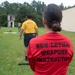 An outdoor military training exercise is taking place on a grassy field adjacent to a building. A person wearing a red shirt labeled 'Non-Lethal Weapons Instructor' observes a Navy personnel in a yellow 'NAVY' shirt conducting or participating in training. Additional personnel are visible in the background near what appears to be a training structure.