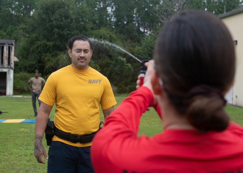 A U.S. Navy service member in a yellow 'NAVY' t-shirt is being sprayed in the face with what appears to be OC (oleoresin capsicum/pepper) spray by a person in a red long-sleeve shirt. This appears to be a military or law enforcement training exercise where personnel are required to be exposed to pepper spray as part of certification. A third individual in the background observes the exercise on a grassy outdoor training area.
