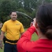 A U.S. Navy service member in a yellow 'NAVY' t-shirt is being sprayed in the face with what appears to be OC (oleoresin capsicum/pepper) spray by a person in a red long-sleeve shirt. This appears to be a military or law enforcement training exercise where personnel are required to be exposed to pepper spray as part of certification. A third individual in the background observes the exercise on a grassy outdoor training area.