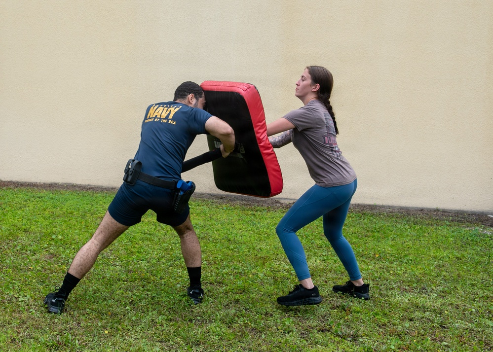 Two individuals are engaged in a martial arts or self-defense training exercise outdoors on a grass lawn. One person wearing a U.S. Navy t-shirt holds a large red and black striking shield while the other person, a woman in a gray shirt and teal leggings, pushes or strikes against it. The setting appears to be near a building with a light-colored stucco wall.