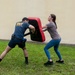 Two individuals are engaged in a martial arts or self-defense training exercise outdoors on a grass lawn. One person wearing a U.S. Navy t-shirt holds a large red and black striking shield while the other person, a woman in a gray shirt and teal leggings, pushes or strikes against it. The setting appears to be near a building with a light-colored stucco wall.