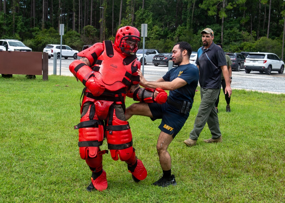 A U.S. Navy service member in a dark blue 'NAVY' t-shirt and shorts is engaged in a combatives or self-defense training exercise on a grass field. He is striking or grappling with an instructor wearing full red padded protective 'redman' suit armor, including helmet, chest guard, and limb padding. A third individual in civilian attire appears to be observing or supervising the drill in the background.