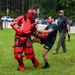 A U.S. Navy service member in a dark blue 'NAVY' t-shirt and shorts is engaged in a combatives or self-defense training exercise on a grass field. He is striking or grappling with an instructor wearing full red padded protective 'redman' suit armor, including helmet, chest guard, and limb padding. A third individual in civilian attire appears to be observing or supervising the drill in the background.