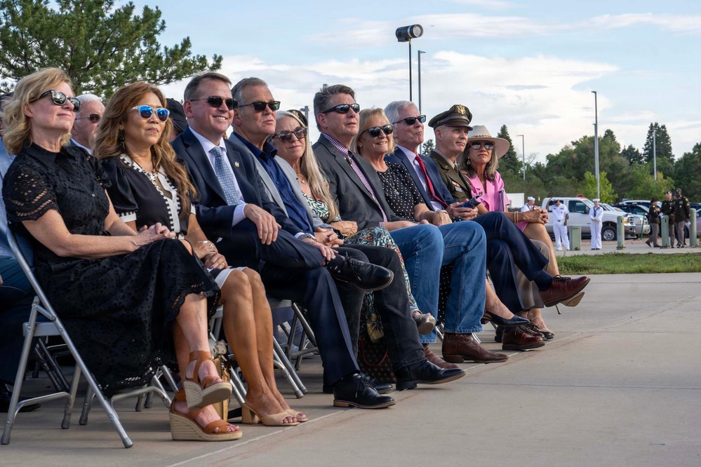 A group of approximately 12 adults are seated in folding chairs outdoors, appearing to attend a formal ceremony or official event. Most individuals are wearing sunglasses and business or semi-formal attire, with one person visible in a military dress uniform. In the background, uniformed military personnel in white dress uniforms and law enforcement officers are visible near vehicles.