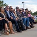 A group of approximately 12 adults are seated in folding chairs outdoors, appearing to attend a formal ceremony or official event. Most individuals are wearing sunglasses and business or semi-formal attire, with one person visible in a military dress uniform. In the background, uniformed military personnel in white dress uniforms and law enforcement officers are visible near vehicles.