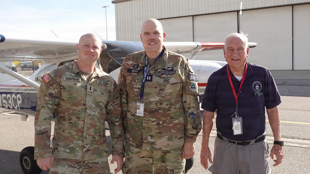Three men pose in front of a small civilian aircraft on an airport tarmac. Two are wearing U.S. military camouflage uniforms, and one is in civilian attire with a lanyard and badge. Name tapes and patches indicate U.S. Army and Civil Air Patrol affiliations.