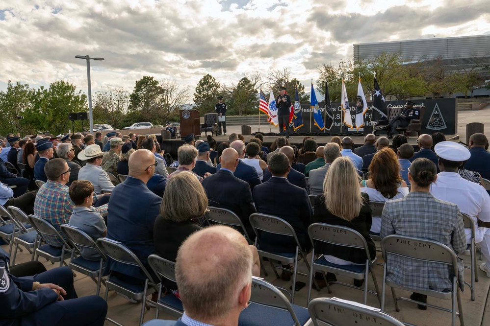 An outdoor military ceremony is taking place with a large audience seated in folding chairs facing a stage. A speaker in military uniform addresses the crowd at a podium, with multiple military and national flags displayed on stage. A banner in the background references 'Headquarters Command' and 'U.S. Space Force' with mention of a 'Hartman Building' dedication or commissioning.