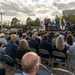 An outdoor military ceremony is taking place with a large audience seated in folding chairs facing a stage. A speaker in military uniform addresses the crowd at a podium, with multiple military and national flags displayed on stage. A banner in the background references 'Headquarters Command' and 'U.S. Space Force' with mention of a 'Hartman Building' dedication or commissioning.