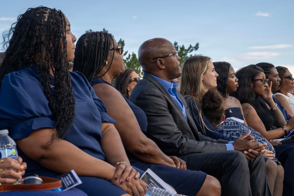 A group of people are seated outdoors in rows, appearing to attend a formal ceremony or memorial service. Most attendees are dressed in navy blue and formal attire, suggesting a coordinated or official event. The gathering appears solemn and attentive, with participants focused toward an unseen focal point.