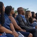 A group of people are seated outdoors in rows, appearing to attend a formal ceremony or memorial service. Most attendees are dressed in navy blue and formal attire, suggesting a coordinated or official event. The gathering appears solemn and attentive, with participants focused toward an unseen focal point.