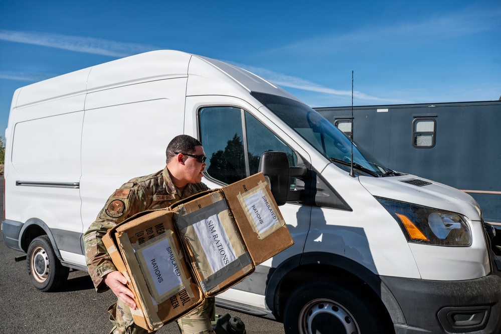 A military service member in OCP (Operational Camouflage Pattern) uniform is carrying multiple cardboard boxes labeled 'SIMULATED RATIONS' near a white cargo van. The scene appears to be a training or logistics exercise, as indicated by the 'simulated' labeling on the boxes. The setting is an outdoor paved area under clear blue skies.
