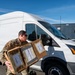 A military service member in OCP (Operational Camouflage Pattern) uniform is carrying multiple cardboard boxes labeled 'SIMULATED RATIONS' near a white cargo van. The scene appears to be a training or logistics exercise, as indicated by the 'simulated' labeling on the boxes. The setting is an outdoor paved area under clear blue skies.