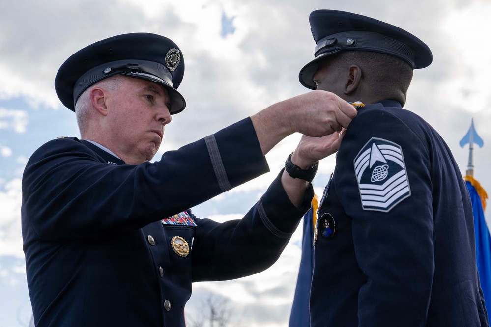 Two military personnel in formal dress uniforms are participating in a medal or decoration ceremony outdoors. A senior officer on the left is pinning a medal or award onto the uniform of another service member on the right, who displays a Chief Master Sergeant rank insignia. The ceremony appears to be taking place against an overcast sky with a military flag visible in the background.