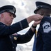 Two military personnel in formal dress uniforms are participating in a medal or decoration ceremony outdoors. A senior officer on the left is pinning a medal or award onto the uniform of another service member on the right, who displays a Chief Master Sergeant rank insignia. The ceremony appears to be taking place against an overcast sky with a military flag visible in the background.