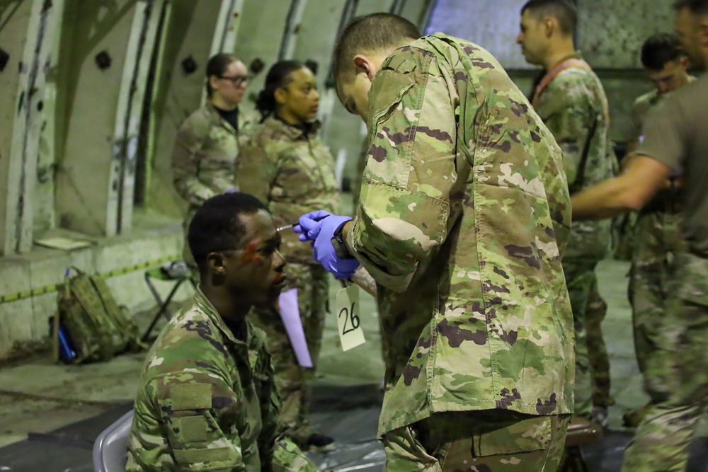 Military personnel in MultiCam/OCP camouflage uniforms are conducting what appears to be a medical training exercise inside a large military aircraft (likely a C-17 or C-5). A standing soldier wearing blue medical gloves is attending to a seated soldier who has simulated wound makeup (red marks) on his face and is tagged with a triage number '26'. Several other uniformed personnel observe or participate in the background.