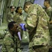 Military personnel in MultiCam/OCP camouflage uniforms are conducting what appears to be a medical training exercise inside a large military aircraft (likely a C-17 or C-5). A standing soldier wearing blue medical gloves is attending to a seated soldier who has simulated wound makeup (red marks) on his face and is tagged with a triage number '26'. Several other uniformed personnel observe or participate in the background.