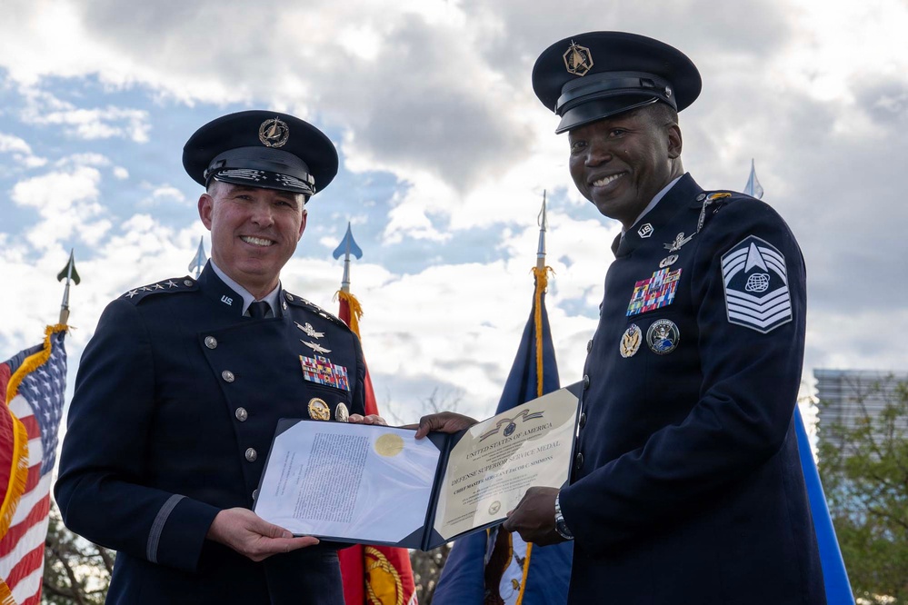 Two U.S. military personnel in dress uniforms are jointly holding an official certificate during what appears to be a formal awards ceremony held outdoors. The document is visibly titled 'Defense Superior Service Medal' issued by the United States of America. Both individuals are smiling, suggesting a celebratory and ceremonial occasion.