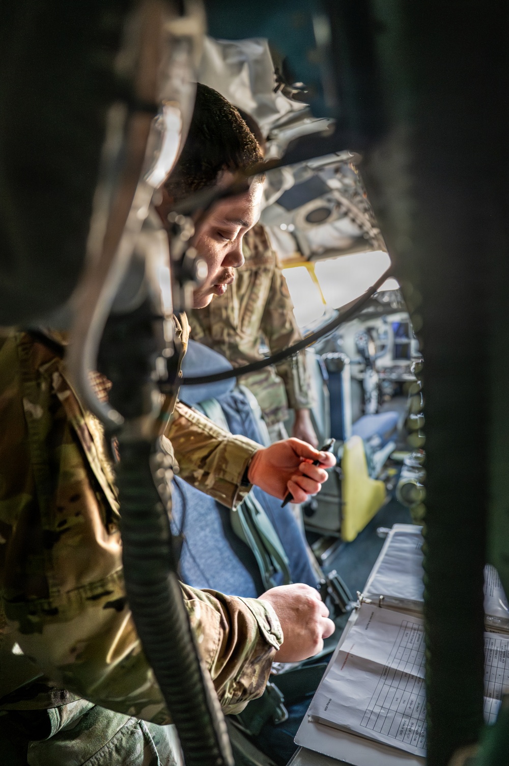 Two military personnel in camouflage uniforms are photographed inside what appears to be a military aircraft cockpit or crew station. The image is taken through structural framework, giving a partially obstructed view. One individual is holding a pen and appears to be reviewing documents in a binder, suggesting a pre-flight checklist or inspection procedure.