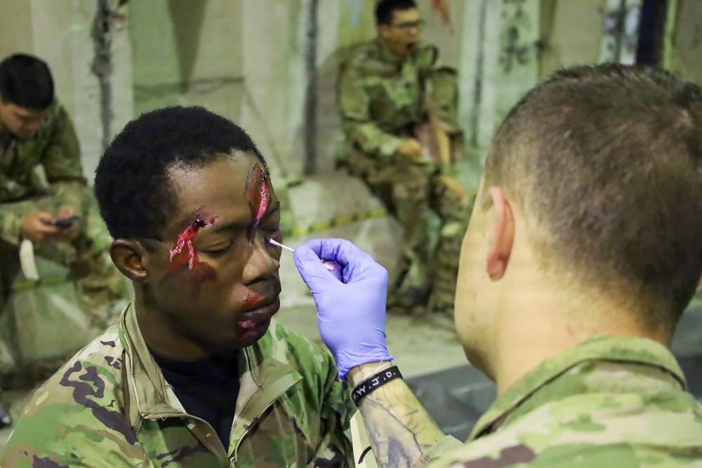 A military training exercise involving simulated casualty makeup application. A soldier wearing blue latex gloves is applying simulated wound makeup (fake blood) to another soldier's face using a cotton swab. Additional soldiers in camouflage uniforms are visible in the background, suggesting a mass casualty training scenario.