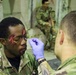 A military training exercise involving simulated casualty makeup application. A soldier wearing blue latex gloves is applying simulated wound makeup (fake blood) to another soldier's face using a cotton swab. Additional soldiers in camouflage uniforms are visible in the background, suggesting a mass casualty training scenario.