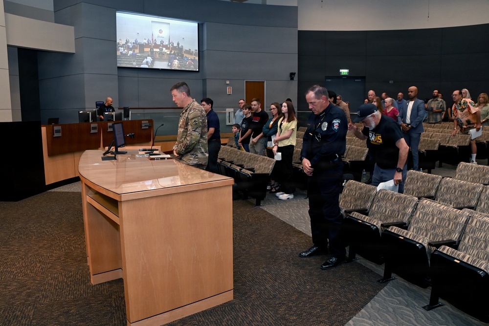 A formal public meeting or ceremony is taking place in what appears to be a government council chamber or civic auditorium. A military service member in camouflage uniform and a police officer in dark uniform are standing near a podium, with a crowd of attendees standing in the background. A large display screen above shows what appears to be a live feed of a council or board meeting.