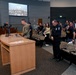 A formal public meeting or ceremony is taking place in what appears to be a government council chamber or civic auditorium. A military service member in camouflage uniform and a police officer in dark uniform are standing near a podium, with a crowd of attendees standing in the background. A large display screen above shows what appears to be a live feed of a council or board meeting.