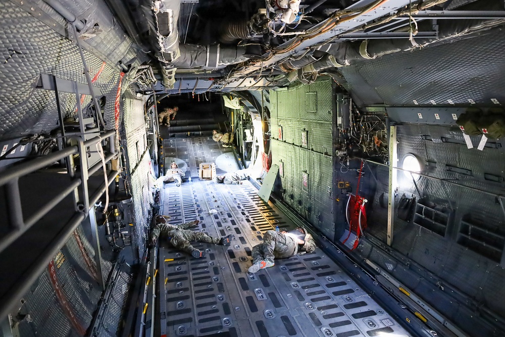 The image shows the interior of a large military transport aircraft, likely a C-130 Hercules or similar cargo plane, with several military personnel lying on the cargo floor. The wide-angle perspective reveals the full length of the aircraft's cargo bay, including exposed structural elements, wiring, and tie-down tracks on the floor. Personnel in military camouflage uniforms are positioned at various points along the floor, possibly resting during transport.