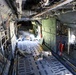 The image shows the interior of a large military transport aircraft, likely a C-130 Hercules or similar cargo plane, with several military personnel lying on the cargo floor. The wide-angle perspective reveals the full length of the aircraft's cargo bay, including exposed structural elements, wiring, and tie-down tracks on the floor. Personnel in military camouflage uniforms are positioned at various points along the floor, possibly resting during transport.