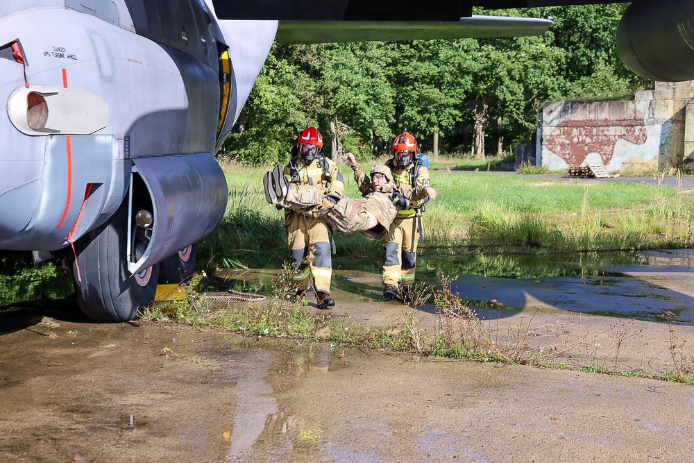 Two firefighters in full protective gear and red helmets are carrying an incapacitated person (likely a simulated casualty in military-style clothing) away from a large military aircraft during what appears to be an emergency response training exercise. The scene takes place on a wet, deteriorated airfield tarmac beside the landing gear of a gray military transport aircraft. The surrounding environment includes overgrown grass, standing water, and a dilapidated structure in the background, suggesting an abandoned or decommissioned airfield used for training purposes.