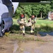 Two firefighters in full protective gear and red helmets are carrying an incapacitated person (likely a simulated casualty in military-style clothing) away from a large military aircraft during what appears to be an emergency response training exercise. The scene takes place on a wet, deteriorated airfield tarmac beside the landing gear of a gray military transport aircraft. The surrounding environment includes overgrown grass, standing water, and a dilapidated structure in the background, suggesting an abandoned or decommissioned airfield used for training purposes.