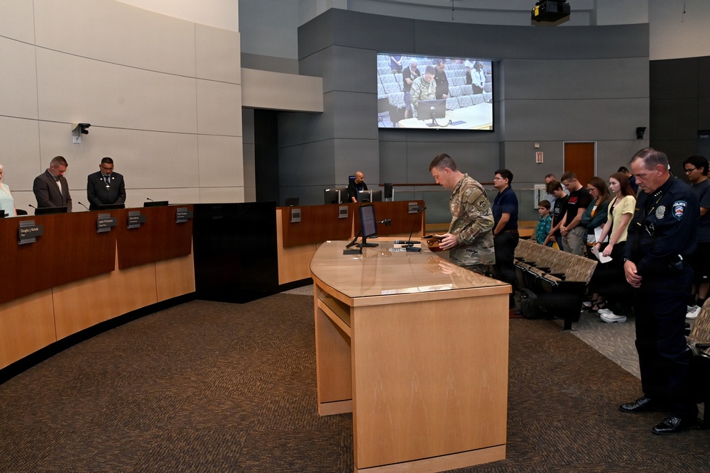 A formal government or civic meeting is taking place in what appears to be a city council chamber. A person in military camouflage uniform stands at a podium, while attendees including a police officer, council members, and civilians bow their heads, suggesting a moment of prayer or invocation. The scene is being recorded and displayed on a large screen at the back of the room.