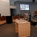 A formal government or civic meeting is taking place in what appears to be a city council chamber. A person in military camouflage uniform stands at a podium, while attendees including a police officer, council members, and civilians bow their heads, suggesting a moment of prayer or invocation. The scene is being recorded and displayed on a large screen at the back of the room.