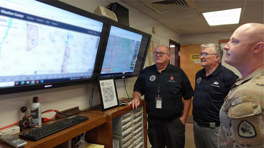 Three individuals are gathered in what appears to be an emergency operations or weather monitoring center, reviewing geographic mapping data displayed on two large Sony monitors. Two men in black polo shirts with name tags and one person in military camouflage uniform are observing the screens attentively. The setting suggests a coordinated civil-military emergency management or situational awareness briefing.
