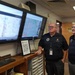 Three individuals are gathered in what appears to be an emergency operations or weather monitoring center, reviewing geographic mapping data displayed on two large Sony monitors. Two men in black polo shirts with name tags and one person in military camouflage uniform are observing the screens attentively. The setting suggests a coordinated civil-military emergency management or situational awareness briefing.