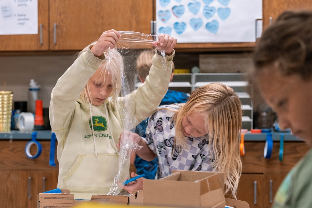 Two young students are engaged in a hands-on classroom activity, working with plastic wrap or a plastic bag and cardboard boxes. The setting appears to be an elementary school classroom or science lab, with supplies and decorations visible in the background. One child is wearing a John Deere hoodie and holding up a clear plastic sheet, while the other examines a cardboard box.