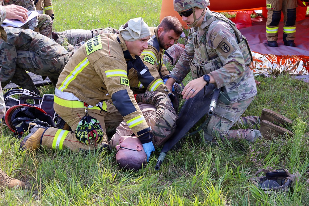 The image depicts a joint military and civilian emergency response training exercise, likely in Poland based on the 'PSP' (Państwowa Straż Pożarna - Polish State Fire Service) markings on the firefighter's uniform. Personnel in military camouflage (OCP pattern) and a firefighter in yellow high-visibility gear are treating a simulated casualty lying on the ground. The scene involves multiple responders coordinating a medical response scenario on a grassy outdoor area.