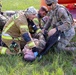 The image depicts a joint military and civilian emergency response training exercise, likely in Poland based on the 'PSP' (Państwowa Straż Pożarna - Polish State Fire Service) markings on the firefighter's uniform. Personnel in military camouflage (OCP pattern) and a firefighter in yellow high-visibility gear are treating a simulated casualty lying on the ground. The scene involves multiple responders coordinating a medical response scenario on a grassy outdoor area.