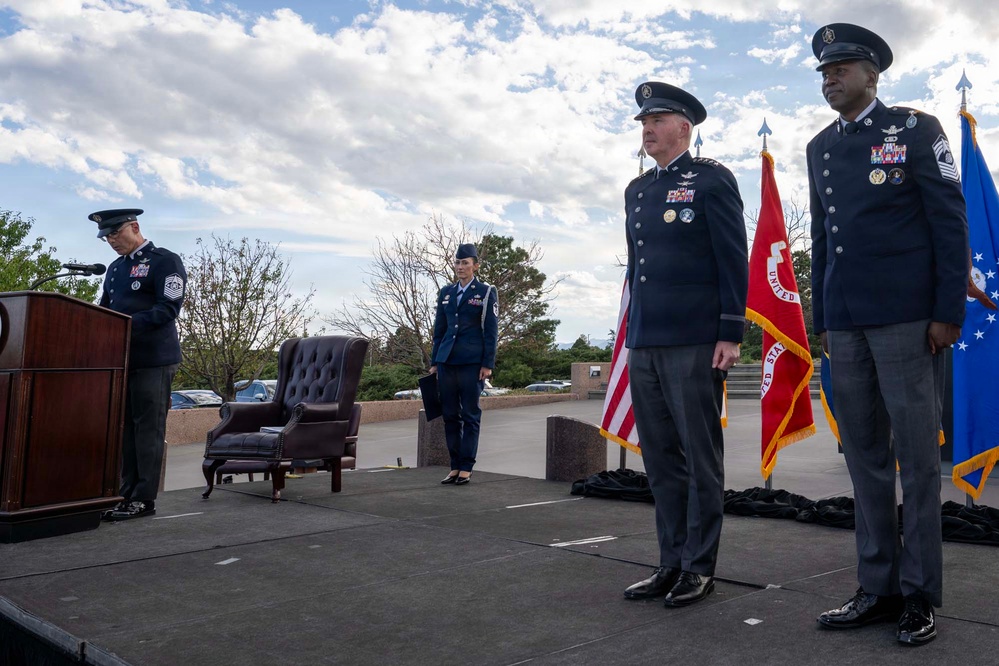A formal U.S. Air Force military ceremony is taking place on an outdoor stage, likely a change of command or retirement ceremony. Several uniformed personnel in dress blues are positioned on the stage, with flags including the U.S. flag and what appears to be a unit guidon visible in the background. One officer is speaking at a podium while others stand at attention.