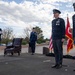 A formal U.S. Air Force military ceremony is taking place on an outdoor stage, likely a change of command or retirement ceremony. Several uniformed personnel in dress blues are positioned on the stage, with flags including the U.S. flag and what appears to be a unit guidon visible in the background. One officer is speaking at a podium while others stand at attention.