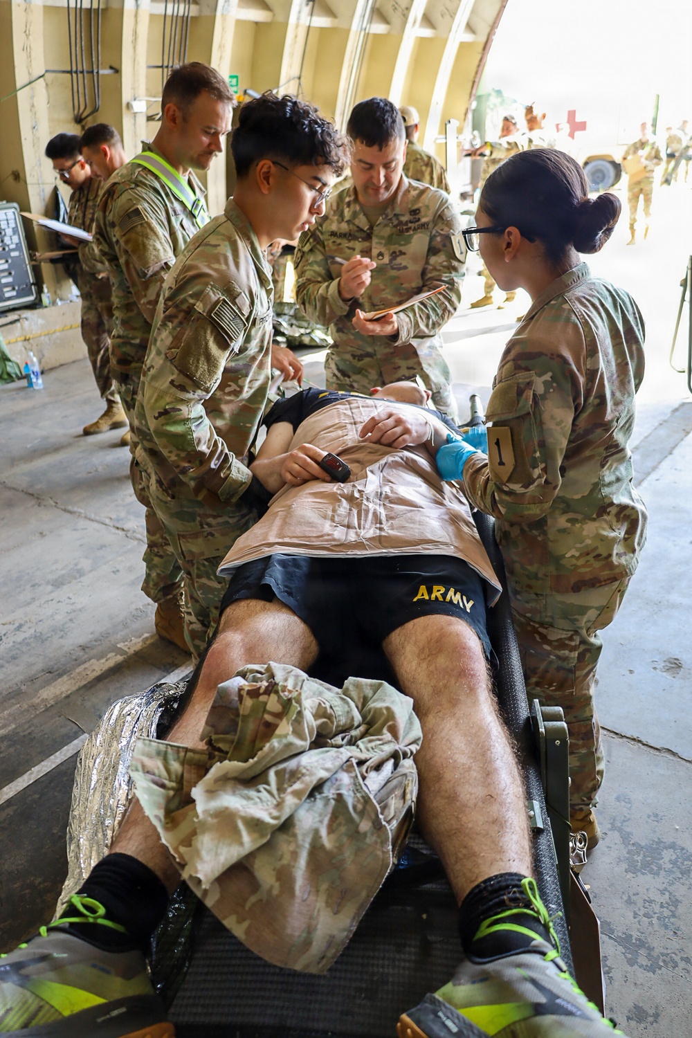 U.S. Army personnel are conducting a medical training exercise, with multiple soldiers in MultiCam uniforms attending to a simulated casualty lying on a litter/stretcher. The patient is wearing Army PT shorts and has simulated wound dressings applied to the lower legs. Several medics are performing assessments while one observer with a yellow vest takes notes, suggesting a graded evaluation scenario.