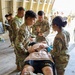 U.S. Army personnel are conducting a medical training exercise, with multiple soldiers in MultiCam uniforms attending to a simulated casualty lying on a litter/stretcher. The patient is wearing Army PT shorts and has simulated wound dressings applied to the lower legs. Several medics are performing assessments while one observer with a yellow vest takes notes, suggesting a graded evaluation scenario.